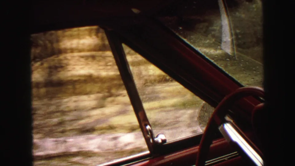The 1967 Yellowstone road-trip view continues through rainy car glass, showing red interior trim, a steering wheel edge, and rocky terrain outside.