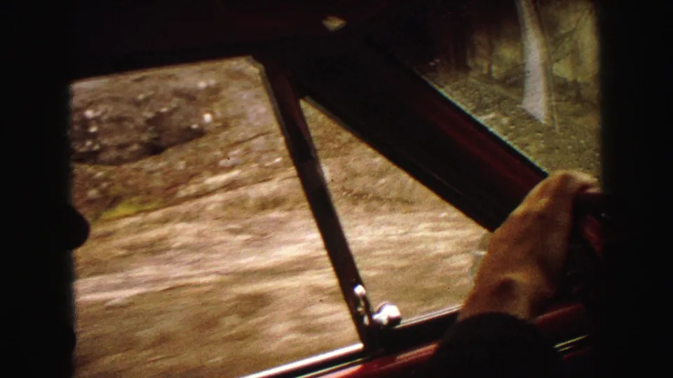 Inside a moving car in the Yellowstone, Wyoming area, a hand holds the wheel while dirt roadside blurs past the window.