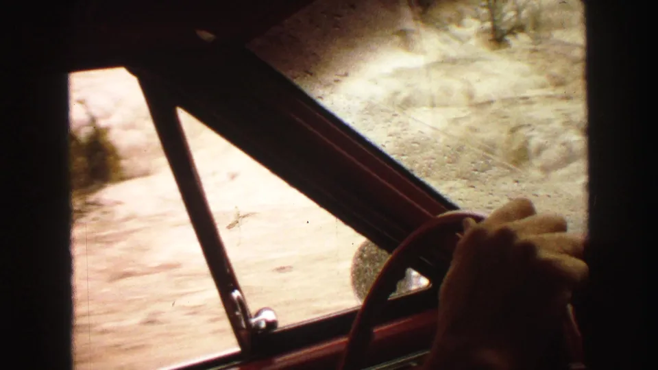 1967 8mm road-trip frame showing a hand near the steering wheel, red window frame, and sunlit rural ground outside.