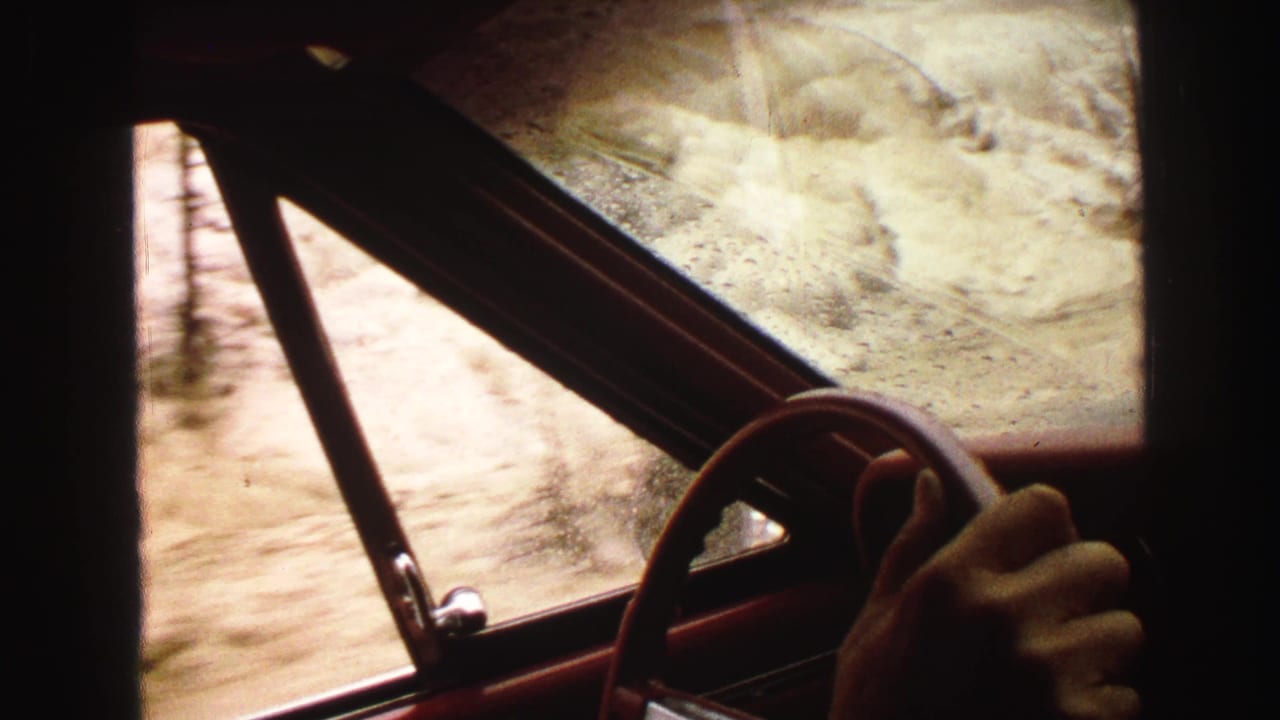View from inside a red car with a hand on the steering wheel beside a wet window and pale dirt road.