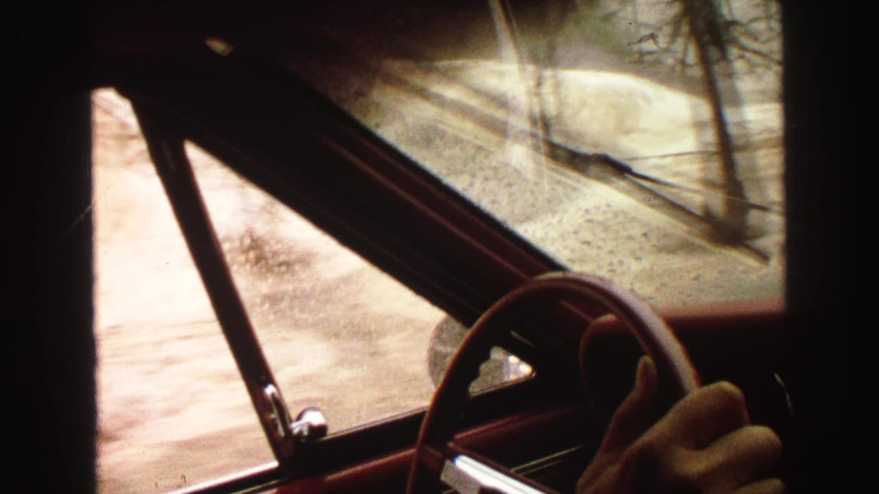 Red car interior with steering wheel and hand visible as wet roadside hills pass outside the window.