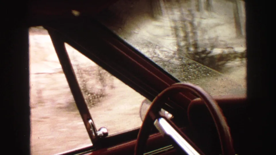 Rainy 8mm driving view from a red car interior in the Yellowstone, Wyoming area, with roadside trees and dirt track outside.