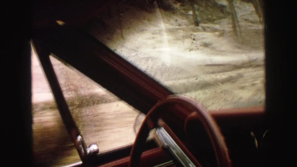 Final frame of a 1967 Yellowstone-area rainy drive, seen from inside a red car with the steering wheel and wet window prominent.
