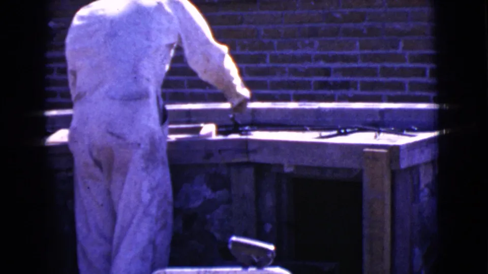 A person in a white jumpsuit works at a wooden counter under purple-blue light in the Cottonwood, Arizona 1968 home-movie clip.