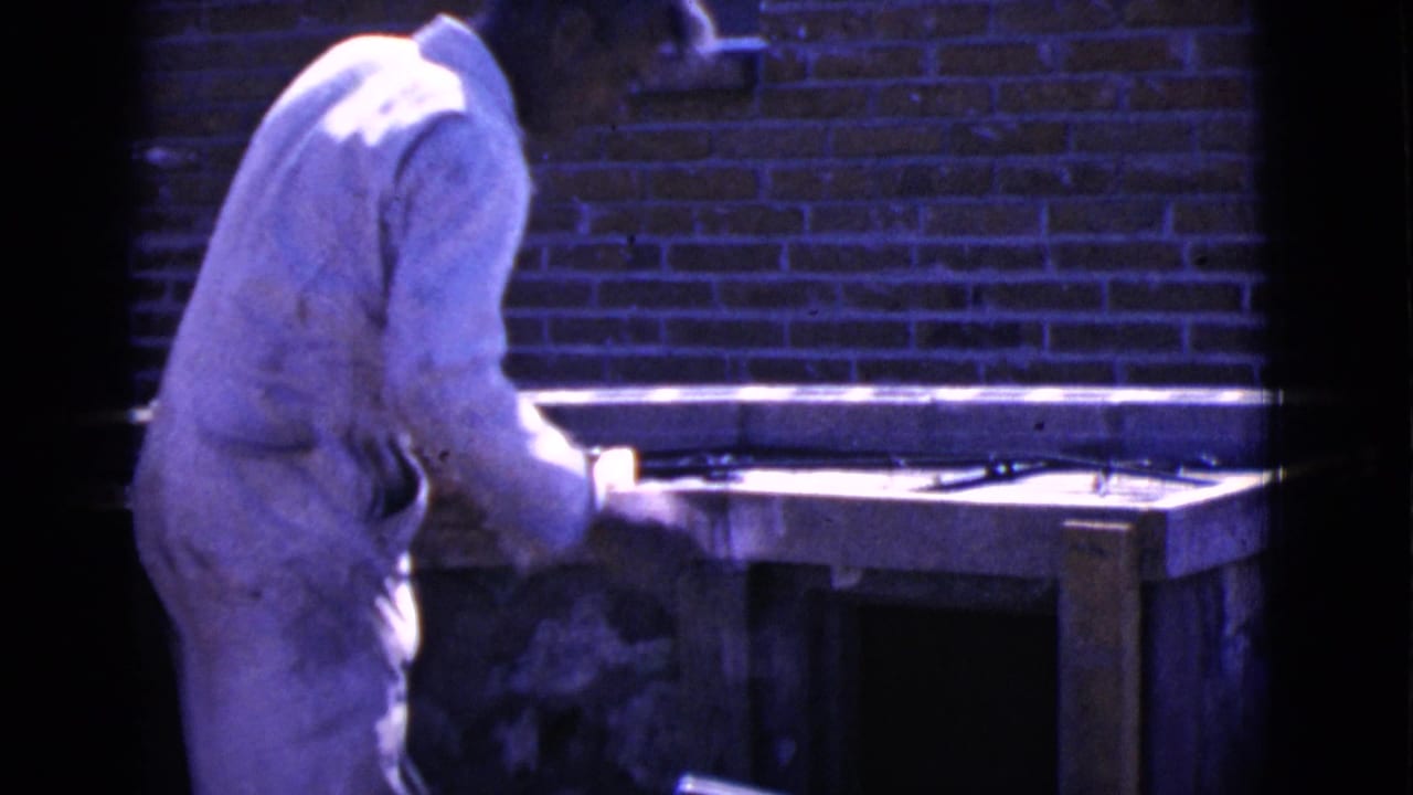 Man in white work clothes stands beside a wood form and mortar near a red brick wall.