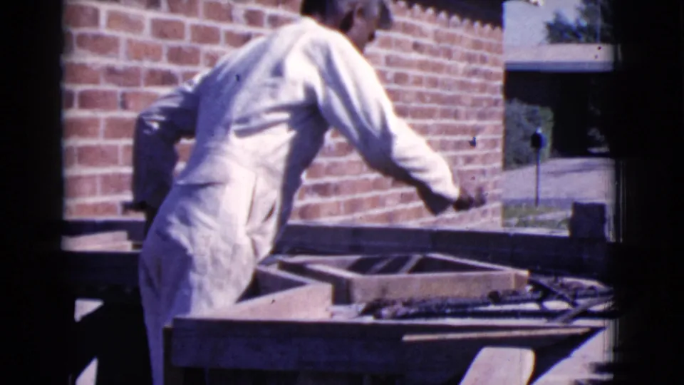 A man in white work clothes reaches over a wooden masonry form outdoors in Cottonwood, Arizona, with a red brick wall behind him.