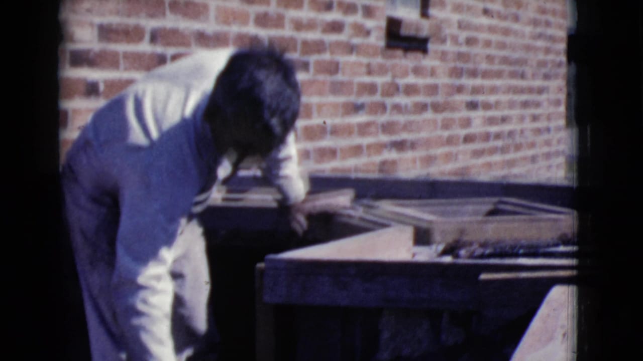 Man in white work clothes bends beside a mortar tray near a red brick wall.