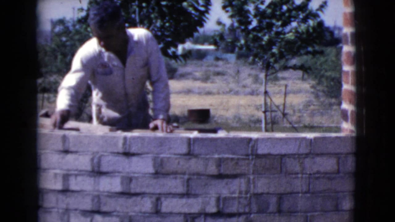 Man leans over a low brick wall while working outdoors.