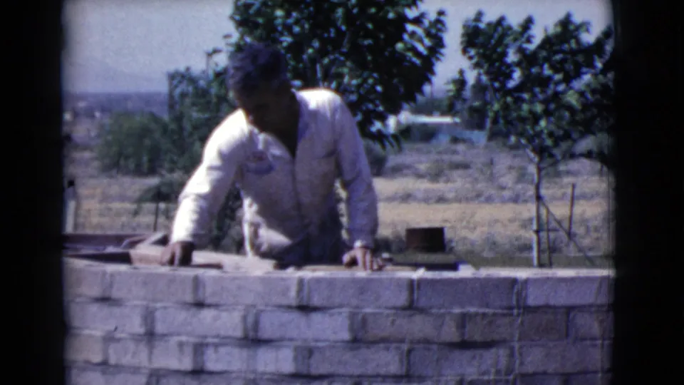 1968 Cottonwood, Arizona home-movie frame showing a man working at a low brick wall, with young trees and open land in the background.