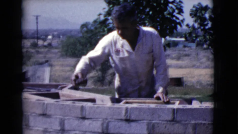 1968 Cottonwood, Arizona frame of a man reaching across a low brick wall during outdoor masonry work, with a dry landscape behind him.