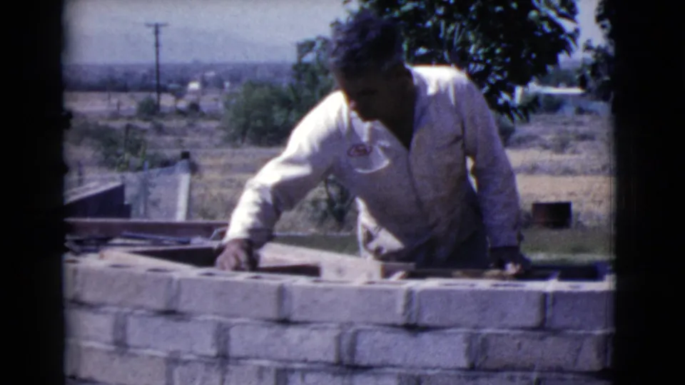 In Cottonwood, Arizona in 1968, a man works on a low concrete block wall in a sunlit yard with trees and distant hills behind him.
