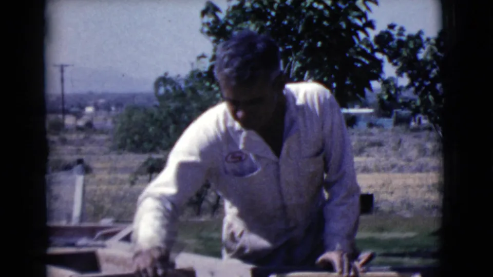 The man leans over a partially built wall, working near wooden forms in the Cottonwood yard captured on 8mm film.