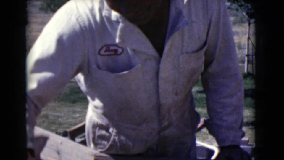 A closer 8mm frame shows the man in a white logo work shirt beside the construction area, with the wall edge and yard partly visible.