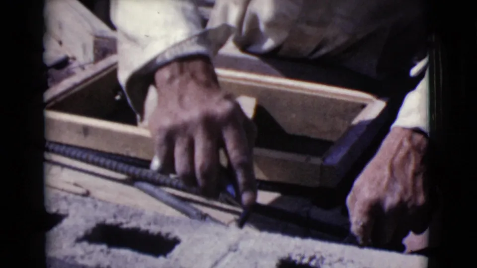 In 1968 Cottonwood, Arizona footage, a worker's hands guide rebar near concrete block cores and wooden form boards.