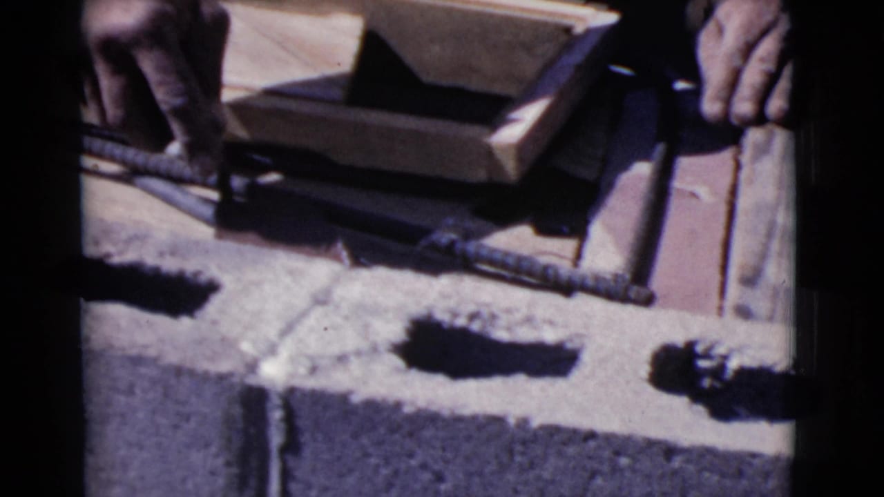 Hands work over concrete blocks with rebar and wooden form boards nearby.