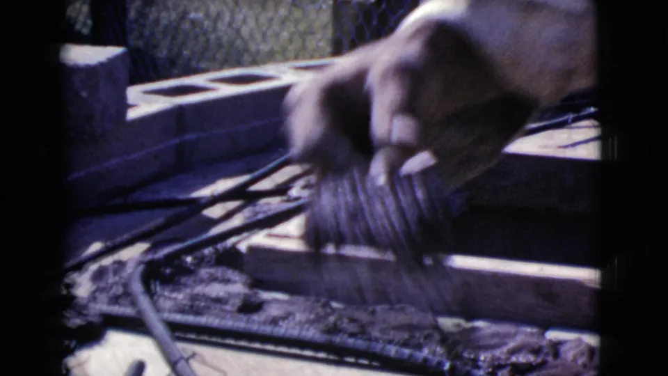 A worker's hand moves above a wooden frame with cinder blocks and rebar visible in bright 1968 Arizona daylight.