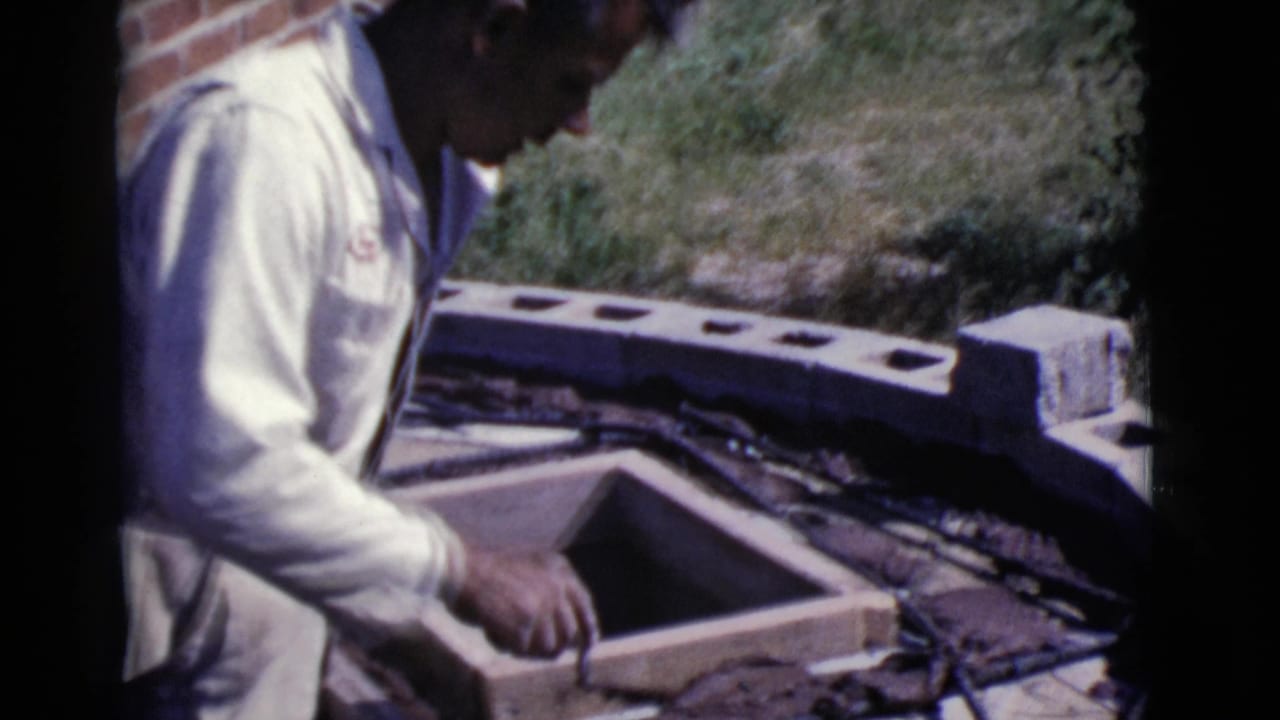Man in white coveralls works beside a wooden form at an outdoor construction area.