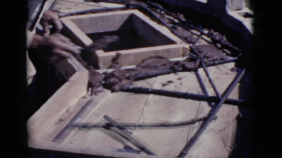 Close view of hands working around a square wooden form with wet grout, rebar, and tools in the Cottonwood construction scene.