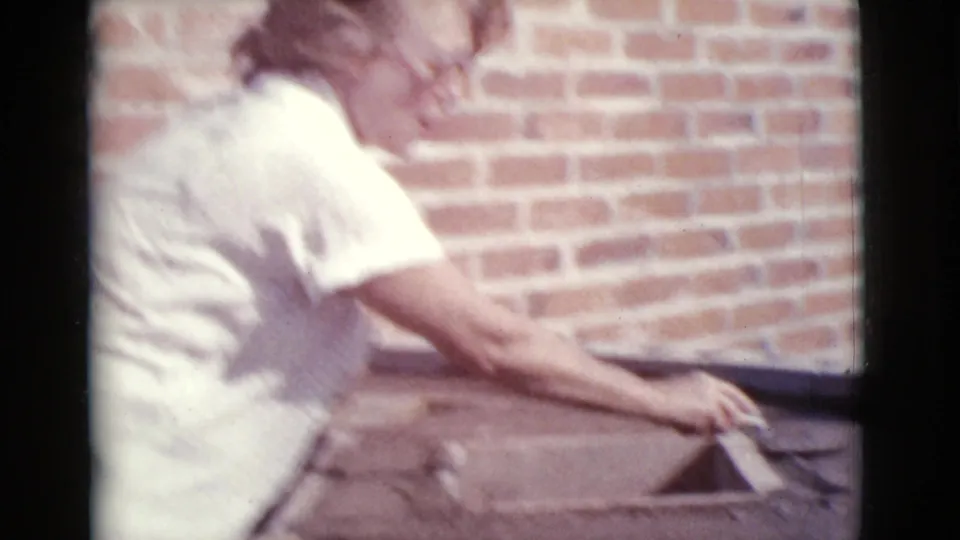 Woman in a light short-sleeved shirt leans over a concrete form beside a brick wall in the 1968 Cottonwood, Arizona home-movie scene.