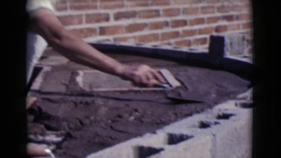 Close 8mm view of a hand troweling wet concrete beside cinder blocks and a brick wall during the Cottonwood, Arizona construction scene.