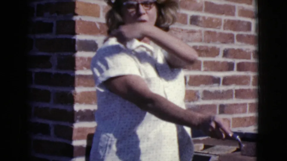 Woman in glasses and a light shirt stands beside a brick wall holding a trowel during the 1968 Cottonwood, Arizona construction footage.