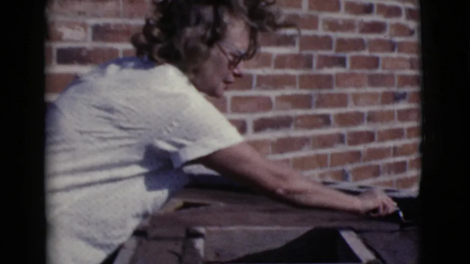 A woman with glasses reaches across a surface beside a red brick wall during outdoor work in Cottonwood, Arizona, 1968.