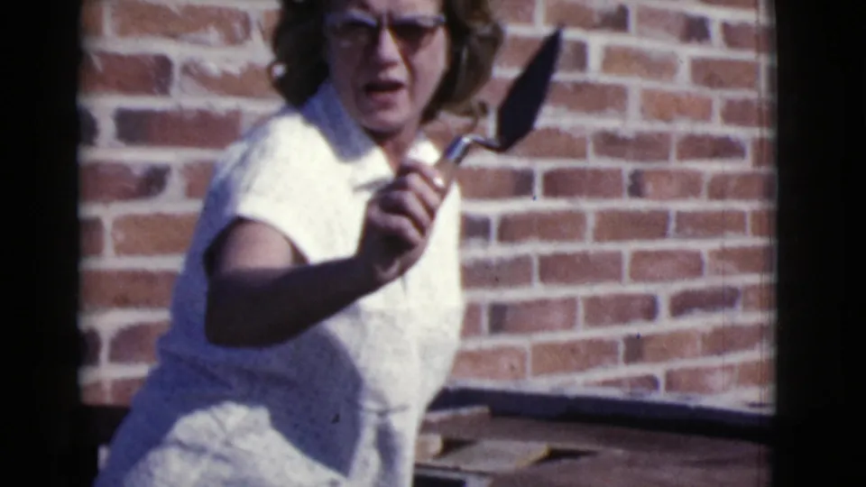 A woman with glasses holds a small trowel toward the camera near a red brick wall in 1968 Cottonwood home-movie footage.