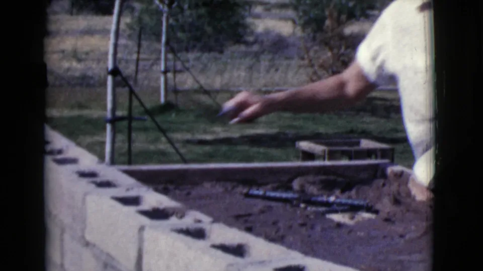 A cropped view shows an arm reaching above a concrete block wall with fresh mortar and yard equipment behind it.
