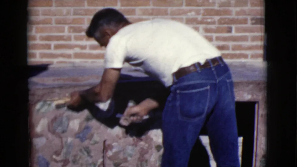 A man in a white T-shirt and blue jeans bends over a stone-and-brick structure, smoothing mortar in the 1968 Arizona footage.