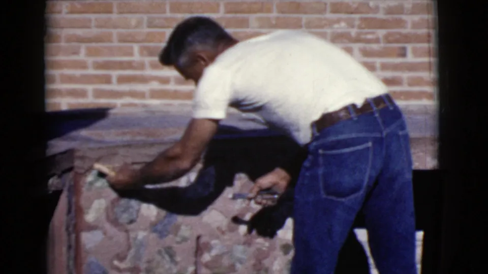 The man continues masonry work on the outdoor stone-and-brick structure, with a brick wall and hard sunlight behind him.