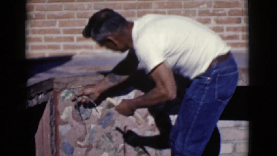 The man continues repairing or adjusting stonework on a fireplace, with a brick wall visible behind him in Cottonwood, Arizona.