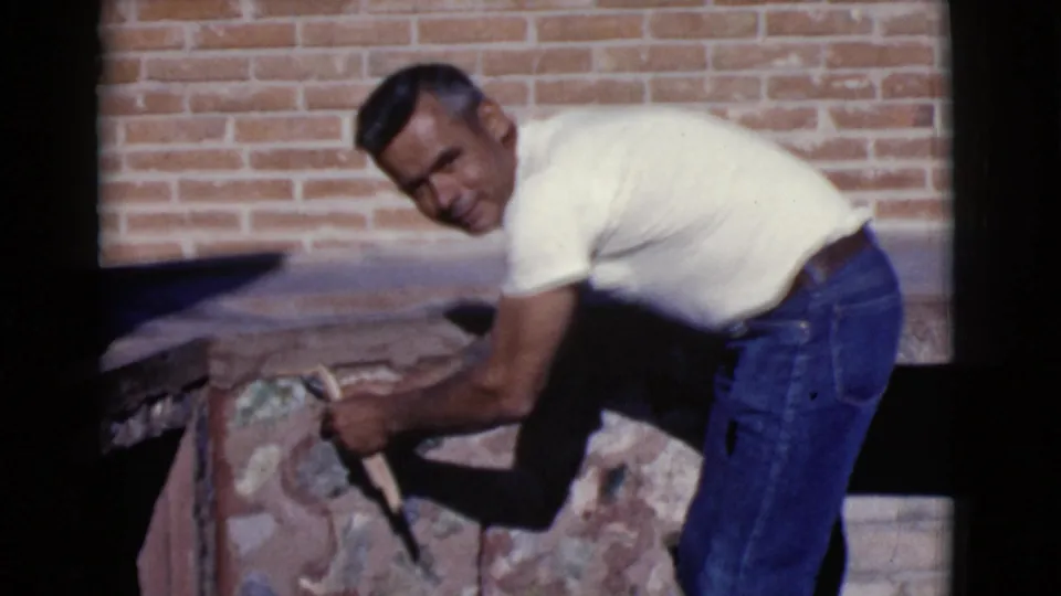 A man in jeans briefly faces the camera while continuing work on a stone fireplace in the 1968 Arizona home-movie scene.