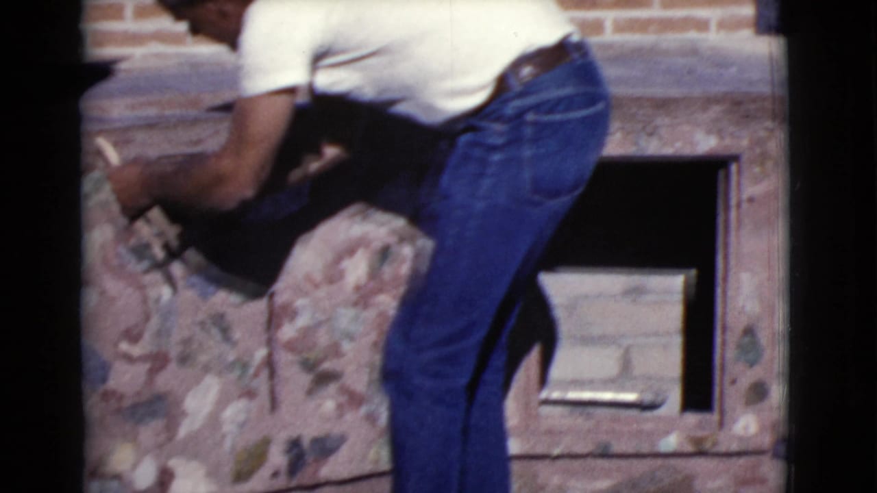 Man in a white shirt and blue jeans works beside a stone structure with a rectangular opening.