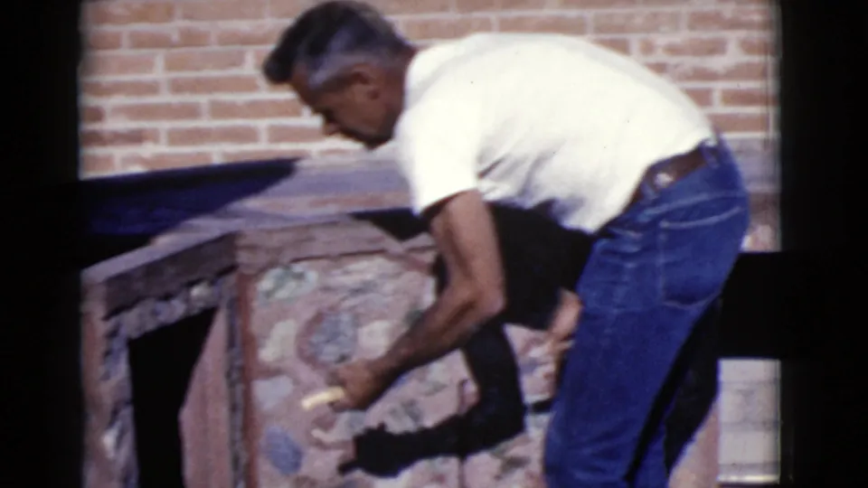 A man in jeans and a white shirt leans over a stone patio feature in Cottonwood, Arizona, with a brick wall behind him.