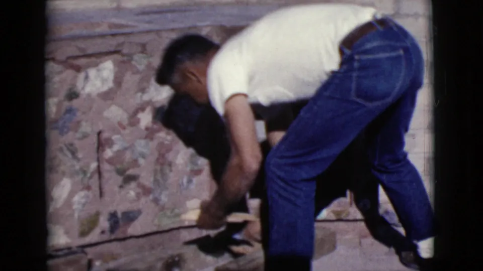 A man in a white shirt and blue jeans bends near the base of a stone patio feature in Cottonwood, Arizona, during a 1968 home movie.