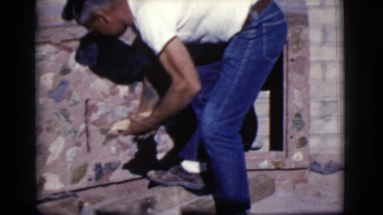 Man in a white shirt and jeans bends near a brick opening while handling material.