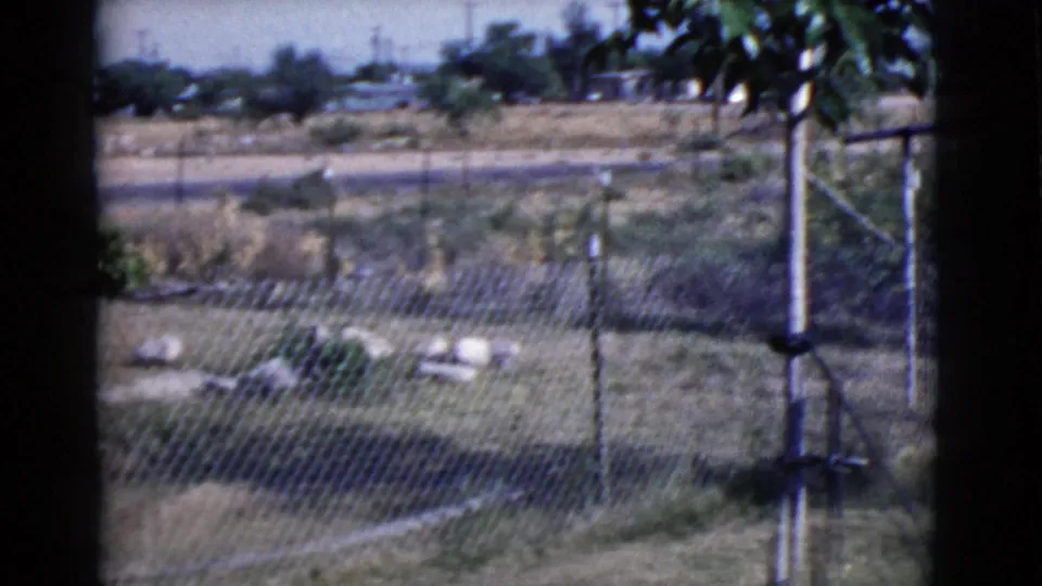 1968 Cottonwood, Arizona 8mm frame showing a quiet fenced yard with dry grass, rocks, a tree trunk, and distant roadside terrain.