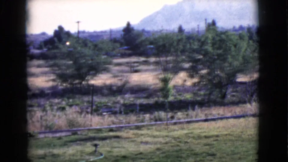 1968 Cottonwood, Arizona landscape with lawn, irrigation hose, trees, fencing, dry field, utility poles, and a hazy mountain beyond.