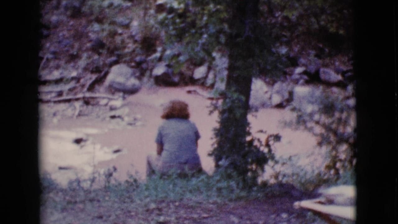 Woman in a blue shirt sits beside muddy water near a tree and rocks.