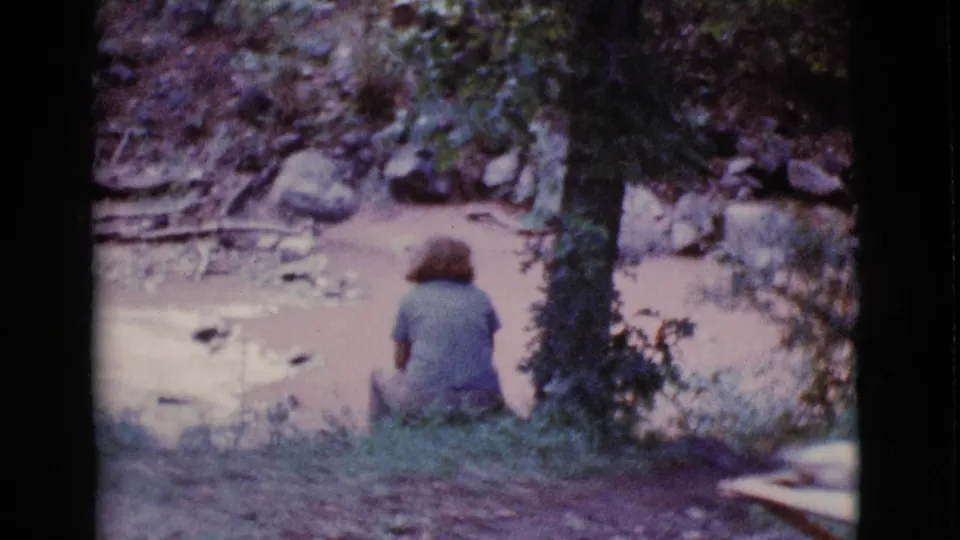 The woman remains seated near a tree beside muddy water in Cottonwood, Arizona, with rocks and low greenery around the bank.