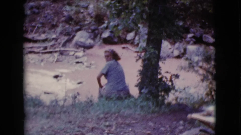 The seated woman turns partway toward the camera while resting beside the muddy pond, with a tree trunk, rocks, and brush visible nearby.