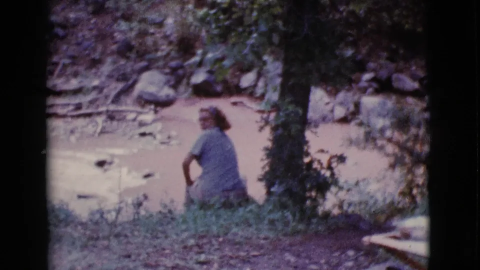 A late-1960s home-movie frame shows the woman in partial profile beside the muddy pond in Cottonwood, Arizona, surrounded by brush, rocks, and shade.