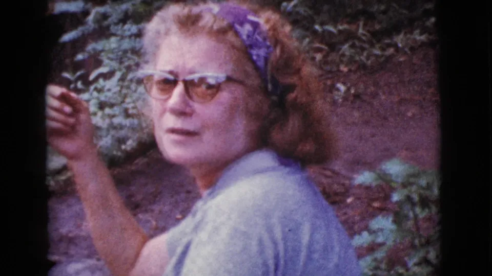 Adult woman wearing glasses and a head scarf sits outdoors in 1968 Arizona home-movie footage, facing the camera.