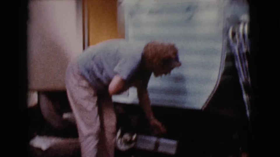Older woman in light clothing bends beside a vintage trailer in Cottonwood, Arizona, 1968, appearing to handle items stored underneath.