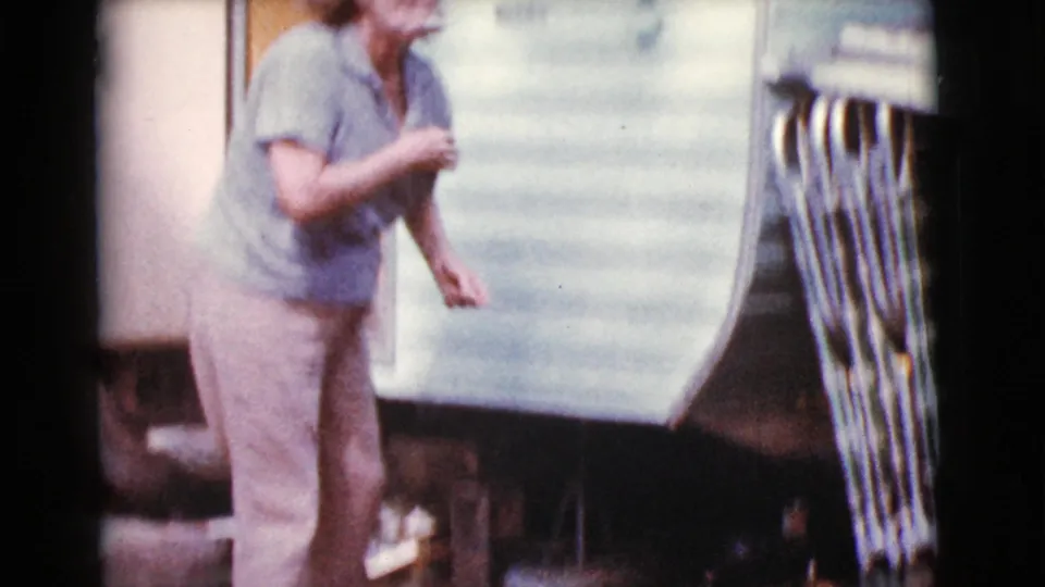 Older woman stands beside a light vintage trailer in Cottonwood, Arizona, 1968, holding a cigarette near camping equipment.