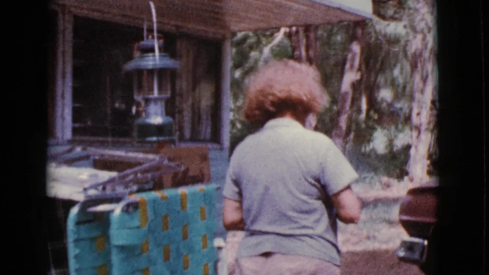 Older woman walks or turns away near a hanging lantern, trailer-side gear, and sunlit outdoor surroundings in the 1968 Cottonwood clip.