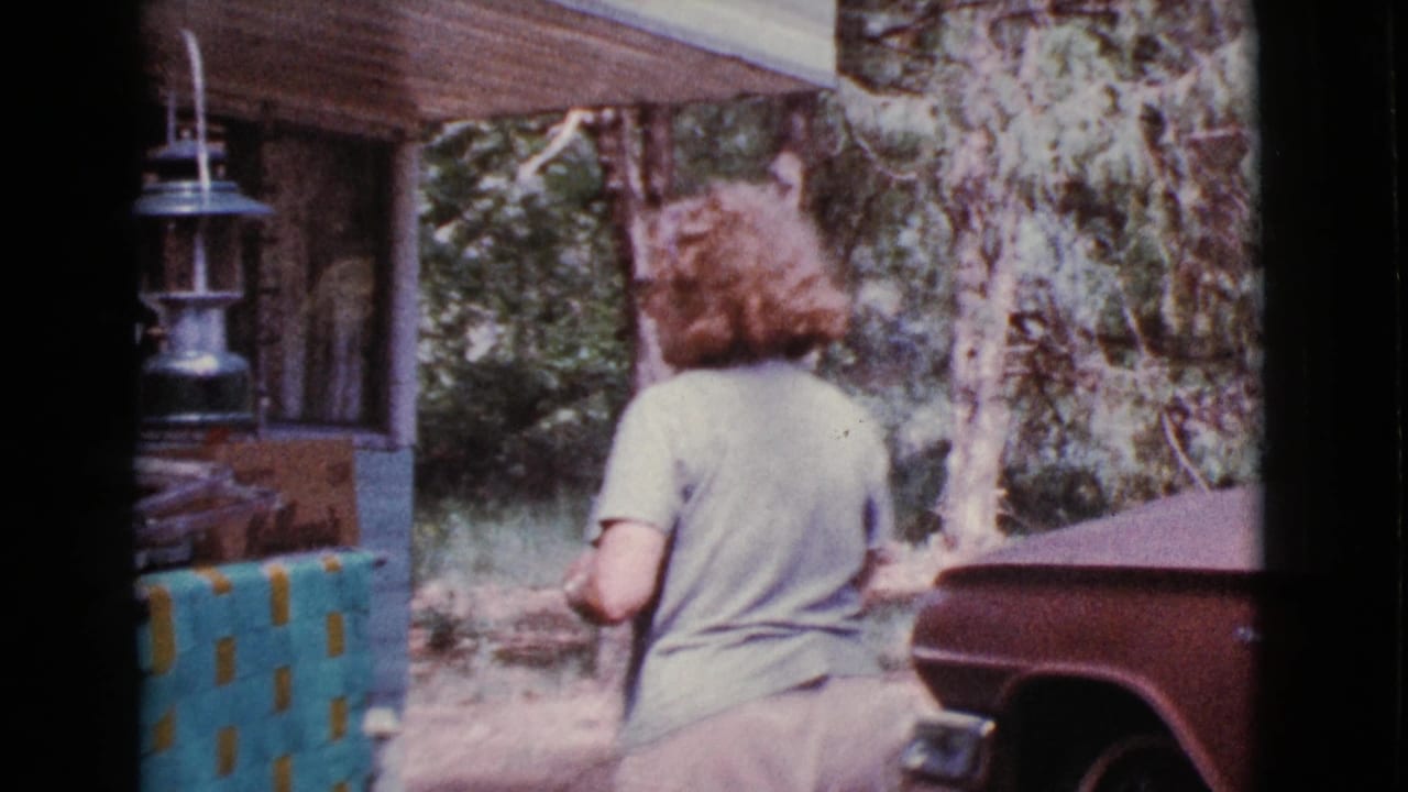 Woman in a light shirt stands beside a trailer porch with a hanging lantern in wooded surroundings.
