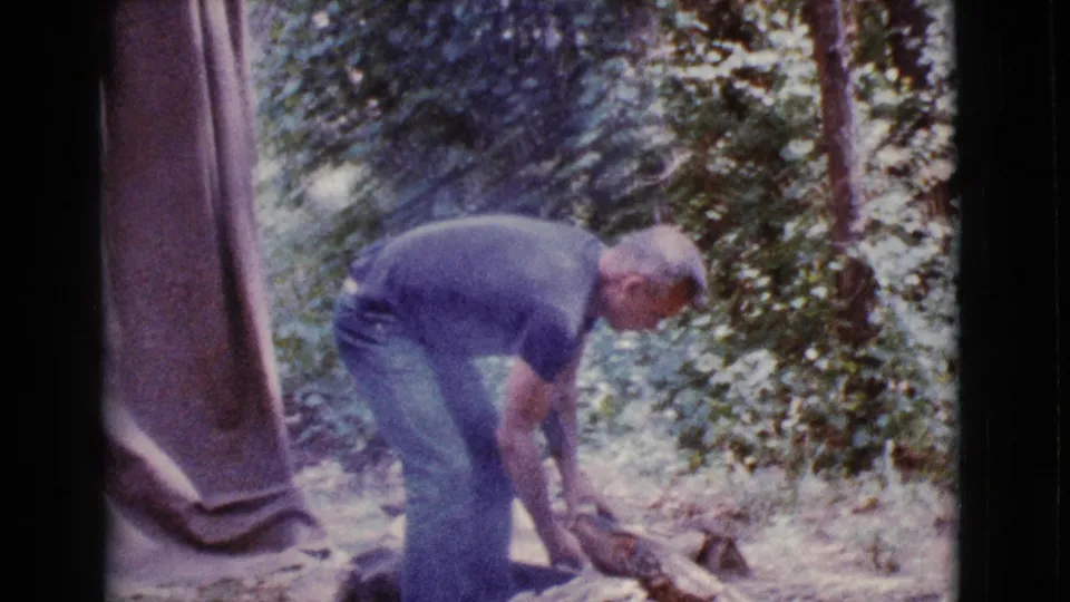 A man bends to gather wood in a wooded Cottonwood, Arizona campsite scene from 1968 8mm home-movie footage.