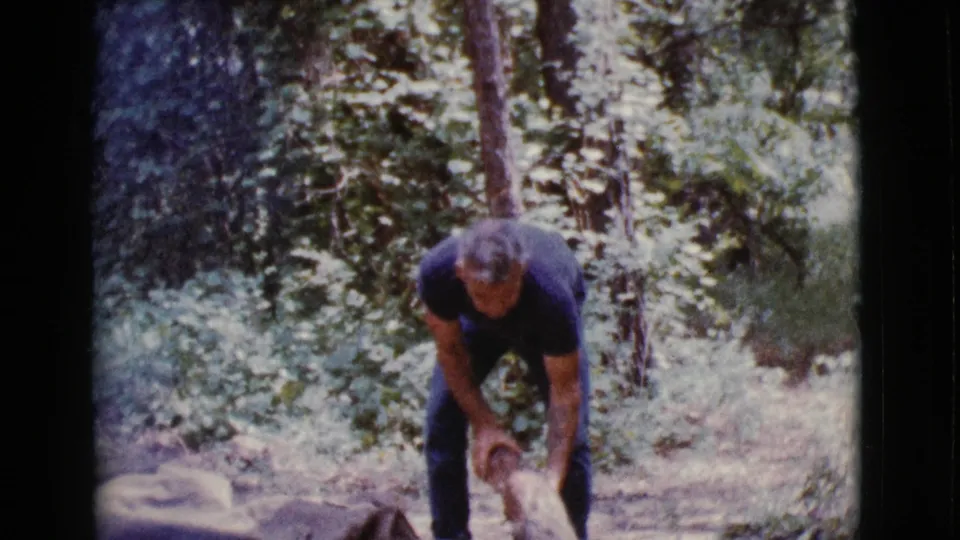 A man bends over with firewood in a wooded Cottonwood, Arizona campsite scene, with soft grain and faded 8mm color.