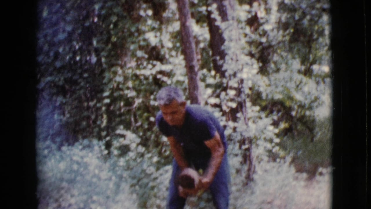 Man bends over a piece of wood in a wooded outdoor area.